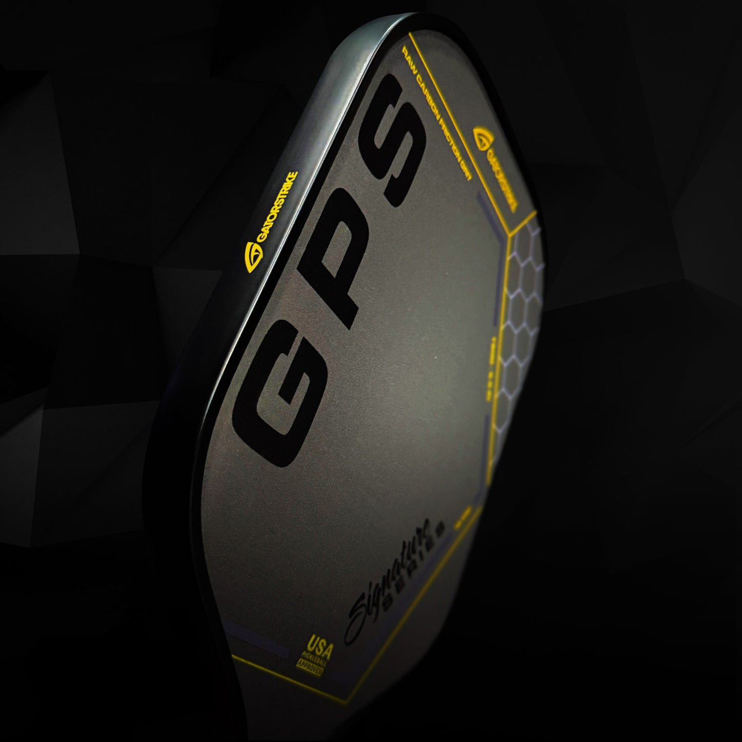 A close-up of the GATORSTRIKE™ Gold Pro Series black carbon fiber paddle, featuring bold GPS lettering and hexagonal yellow accents, displayed against a dark geometric background.